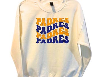 May include: Cream-colored sweatshirt with the word "PADRES" in orange and blue retro font. The word is stacked four times in a wavy design. The sweatshirt has long sleeves and a crew neck.
