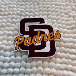 May include: A sticker featuring the San Diego Padres logo. The logo is a stylized "SD" in brown with a white outline, and the word "Padres" in a script font with orange and yellow accents.