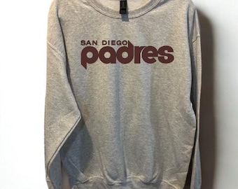 May include: A heather gray sweatshirt with the words "SAN DIEGO Padres" printed in a dark brown color. The sweatshirt is hanging from a black hanger against a white background. The crew neck sweatshirt has long sleeves.