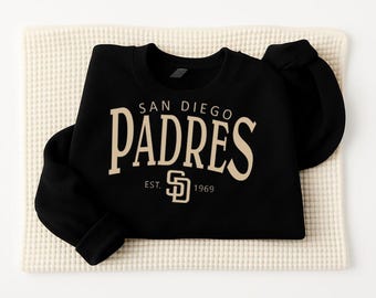May include: Black sweatshirt featuring the words "SAN DIEGO PADRES" in a beige, arched font. Below the team name is "EST. 1969" and the Padres logo. The sweatshirt is folded on a cream-colored, textured surface.