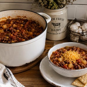May include: A large white enameled cast iron dutch oven filled with hearty chili, accompanied by a matching bowl of chili topped with shredded cheddar cheese and served with square crackers on a plate, featuring a wooden spoon inside the pot.