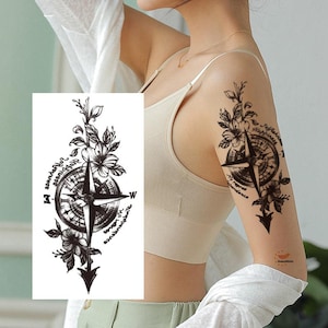 May include: Black temporary tattoo featuring a compass rose design with floral accents and script. The compass is surrounded by flowers and leaves, with the words "Self-reliance, Happiness, No regret, abundance" in a circular pattern.