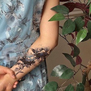 May include: A temporary tattoo featuring a detailed black and grey floral design with shaded peony-like blossoms and delicate stems applied to a forearm. The tattoo is displayed against a light blue dress with a silver floral pattern.
