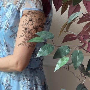 May include: A light blue dress featuring intricate white floral embroidery and a sheer overlay. On the upper arm, a detailed black ink temporary tattoo displays a cluster of lilies and various botanical leaves with fine line work.