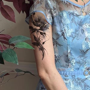 May include: A light blue dress featuring a sheer yoke and intricate silver floral embroidery throughout. The garment has short sleeves and a waistline accented with a circular rhinestone buckle. A black floral temporary tattoo is on the upper arm.