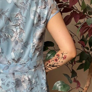 May include: A light blue dress featuring a silver floral embroidery pattern with short sleeves and a defined waistline. The fabric has a sheer overlay with a repeating botanical design. A floral temporary tattoo is visible on the arm.