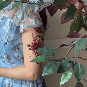 May include: A light blue dress featuring a sheer overlay with intricate silver floral embroidery and short sleeves. A temporary tattoo of dark red and black flowers with thin stems and leaves is applied to the upper arm, showcasing detailed petals.