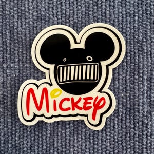 Puede incluir: Una pegatina blanca con un diseño de cabeza de Mickey Mouse negro, con una máscara negra y la palabra "Mickey" en rojo con un punto amarillo en la "i". La pegatina está sobre una superficie azul texturizada.