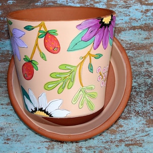 May include: A terracotta flower pot with a hand-painted design featuring pink, purple, white, and green flowers and leaves on a light peach background. The pot has a matching saucer.