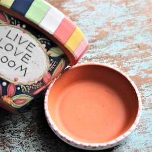 May include: A terracotta flower pot with a colorful striped rim and a painted design featuring flowers and the text "Live Love Bloom". The pot is sitting on a wooden surface next to a matching terracotta saucer.