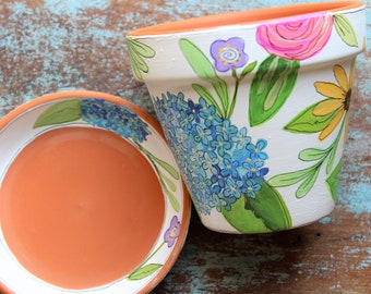 May include: A set of two terracotta flower pots with hand-painted floral designs. The larger pot features blue, yellow, pink, and green flowers, while the smaller pot has a blue and white floral design.