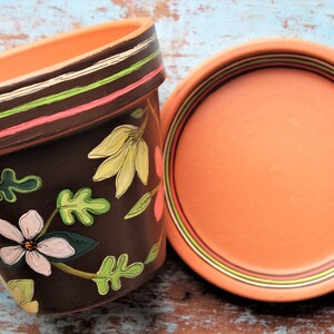 May include: A terracotta flower pot with a brown base and painted floral designs in green, yellow, pink, and white. The pot has a matching saucer with a brown rim and a light orange base.