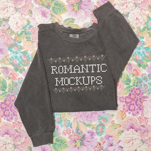 May include: Dark gray sweatshirt with the text "ROMANTIC MOCKUPS" in white, accented with pink floral designs. The sweatshirt is laid on a floral background with pink, purple, and white flowers.