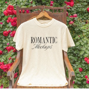 May include: Cream-colored t-shirt with the text "ROMANTIC Mockups" in black script. The shirt hangs on a wooden hanger, set against a backdrop of a wooden chair and vibrant red roses.