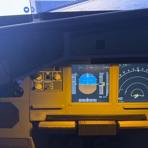 Op de afbeelding: Een close-up van een vliegtuigcockpit met verlichte displays. De schermen tonen vluchtgegevens, waaronder hoogte, snelheid en navigatie-informatie. Verschillende wijzerplaten en schakelaars zijn zichtbaar.