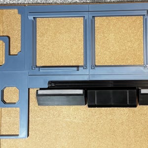 Puede incluir: Un modelo de madera azul grisáceo cortado con láser de un salpicadero de vehículo. El modelo tiene varios recortes para indicadores y controles, con un altavoz negro y una abertura rectangular. El modelo está colocado sobre una superficie marrón claro.