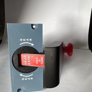 Pode incluir: Um painel retangular cinza com um recorte circular e texto branco escrito GEAR com setas. Uma caixa preta estende-se lateralmente, apresentando uma alavanca vermelha com a inscrição PULL & TURN e GEAR EXTN. Um botão vermelho é visível atrás.