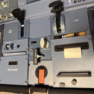 Peut inclure: Une console centrale d'avion grise comprenant un levier de frein aérodynamique, un levier de volets, une poignée de frein de stationnement marquée PARK BRK, une poignée de train d'atterrissage GEAR EXTN, un lecteur de données et une imprimante.