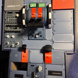May include: A detailed model of an Airbus A320 cockpit panel. Features control levers, switches, and labels such as "REMOVE BEFORE FLIGHT" and "AIRBUS A320". The panel is primarily grey with red and white accents.