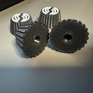 May include: Four black, textured knobs with a ridged edge and a central hole. Two knobs are face up, showing the hole, while the other two are face down, revealing a white design on top. These knobs are likely for electronics or machinery.