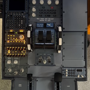 May include: A close-up of an aircraft cockpit control panel, predominantly grey with numerous switches, dials, and displays. Text includes "MAX TAKE OFF WEIGHT REFER TO EFB" and "CABIN SECURE TAKE OFF". The image highlights the intricate nature of flight controls.