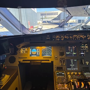 Puede incluir: Vista interior de la cabina de un avión, mostrando una compleja serie de instrumentos, pantallas y controles. La cabina está iluminada con una luz cálida, y el parabrisas ofrece una vista del entorno exterior.