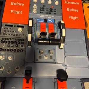 Könnte beinhalten: Flugzeugcockpit-Panel mit verschiedenen Bedienelementen, Schaltern und Hebeln. Zwei orangefarbene Schilder mit der Aufschrift "Remove Before Flight". Das Panel ist grau mit schwarz-weißen Akzenten. Detaillierte Ansicht der Flugzeugsteuerung.