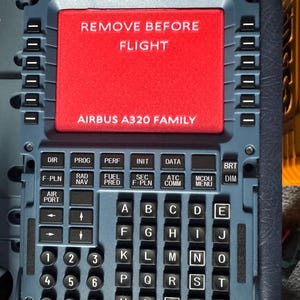 May include: A detailed view of an aircraft control panel. The panel is dark blue with numerous buttons, a keypad, and a red rectangular screen that reads "REMOVE BEFORE FLIGHT" and "AIRBUS A320 FAMILY".