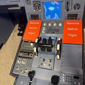 May include: A detailed view of an aircraft cockpit control panel. The panel is primarily gray with various switches, dials, and levers. Two orange signs read "Remove Before Flight." A screen displays a blue water scene.