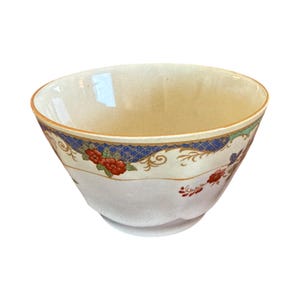 May include: A ceramic bowl with a cream-colored interior and exterior. The upper rim features a decorative band with a blue cross-hatch pattern, red floral motifs, and green leaves. The bowl has a smooth, rounded shape with a subtle scalloped base.