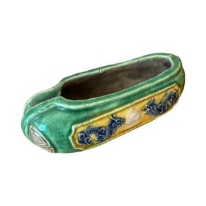 May include: An elongated ceramic vessel with a glossy green glaze and a central yellow panel featuring blue dragon motifs. The piece has a hollow interior, a smooth finish, and a distinct curved shape with a small, textured detail at one end.