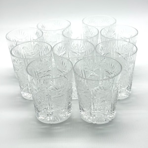 May include: A set of ten clear glass tumblers featuring a pressed geometric starburst and diamond pattern. Each cylindrical glass has a thick base and vertical fluted detailing around the exterior, creating a textured surface that catches light.