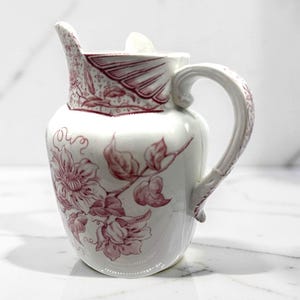 May include: A white ceramic pitcher featuring a detailed maroon floral pattern. The design includes large blossoms and leaves wrapping around the body, with a decorative fan-like motif near the rim and a curved handle with intricate detailing.