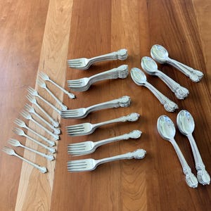 May include: A collection of silver-toned flatware including several forks and spoons. The set features a group of small cocktail forks, medium forks, and spoons with decorative handles. The pieces are arranged in clusters on a wooden surface.