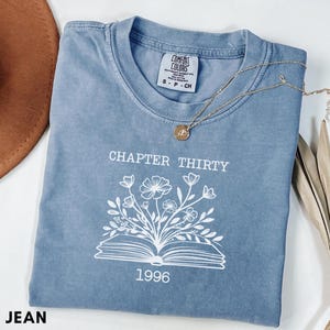 May include: A blue jean Comfort Colors t-shirt with the text "CHAPTER THIRTY" above a floral design and the year "1996". A gold necklace with a pendant rests on the shirt. A brown hat and dried leaves are in the background.