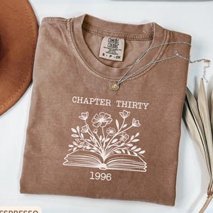 May include: A brown short-sleeve t-shirt featuring a white graphic of an open book with flowers growing out of the pages. The text "CHAPTER THIRTY" is printed above the book and "1996" is printed below. A silver necklace rests on the shirt.