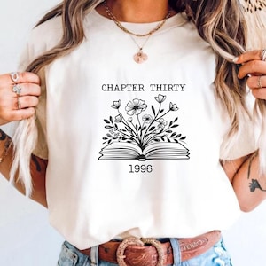 May include: A white t-shirt featuring a black line art graphic of an open book with flowers growing out of the pages. The text "CHAPTER THIRTY" is printed above the book, and the number "1996" is printed below the book in a simple font.