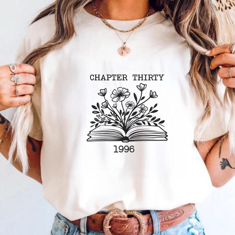 May include: A white t-shirt featuring a black line art graphic of an open book with flowers growing out of the pages. The text "CHAPTER THIRTY" is printed above the book, and the number "1996" is printed below the book in a simple font.