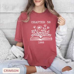 May include: A crimson short-sleeve t-shirt featuring a white graphic of an open book with flowers growing out of the pages. The text "CHAPTER 58" is printed above the book, and the year "1968" is printed below the book in a white font.