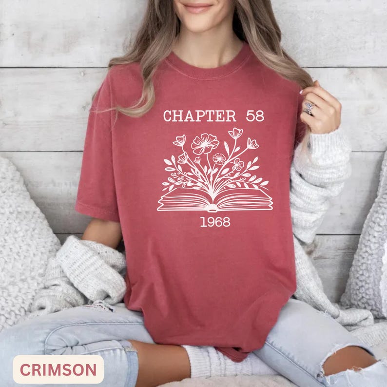 May include: A crimson short-sleeve t-shirt featuring a white graphic of an open book with flowers growing out of the pages. The text "CHAPTER 58" is printed above the book, and the year "1968" is printed below the book in a white font.