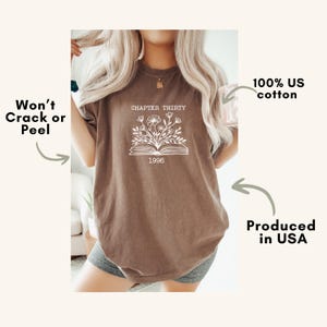 May include: A brown short-sleeve t-shirt featuring a white graphic of an open book with flowers emerging from the pages. The text reads CHAPTER THIRTY and 1996. Additional text on the image states Won't Crack or Peel, 100% US cotton, and Produced in USA.