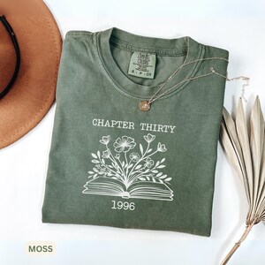 May include: A moss green short-sleeve t-shirt featuring a white graphic of an open book with flowers growing out of the pages. The text "CHAPTER THIRTY" is printed above the book and "1996" is printed below. The shirt has a crew neckline.