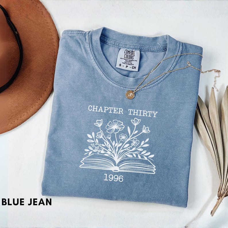 May include: A blue jean Comfort Colors t-shirt with the text "CHAPTER THIRTY" above a floral design and the year "1996". A gold necklace with a pendant rests on the shirt. A brown hat and dried leaves are in the background.