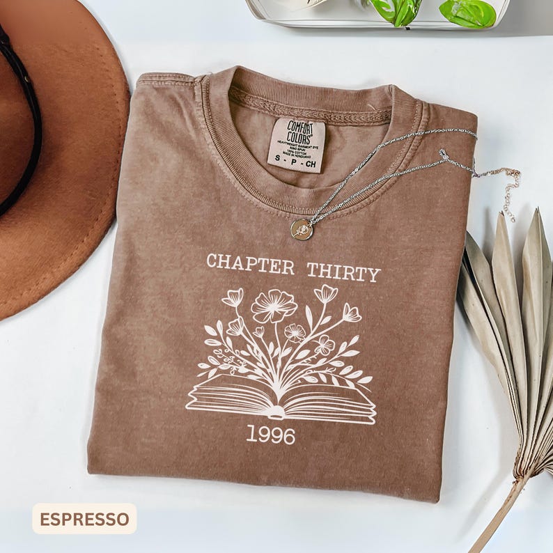 May include: A brown short-sleeve t-shirt featuring a white graphic of an open book with flowers growing out of the pages. The text "CHAPTER THIRTY" is printed above the book and "1996" is printed below. A silver necklace rests on the shirt.