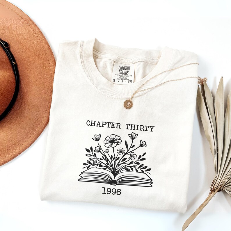 May include: A cream-colored short-sleeve t-shirt featuring a black graphic of an open book with flowers growing out of the pages. The text "CHAPTER THIRTY" is printed above the graphic, and the year "1996" is printed below the book illustration.