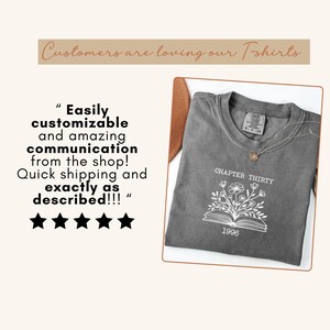 May include: A charcoal gray t-shirt featuring a white line art graphic of an open book with flowers emerging from the pages. The text reads CHAPTER THIRTY and 1996. The shirt has a crew neckline and a visible brand label on the inner collar area.