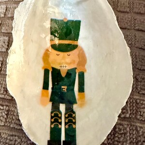 Pode incluir: Enfeite de Natal pintado à mão em forma de concha. O enfeite apresenta um design de soldado quebra-nozes em verde, laranja e preto. Uma fita verde é anexada para pendurar. O fundo é um tecido marrom texturizado.