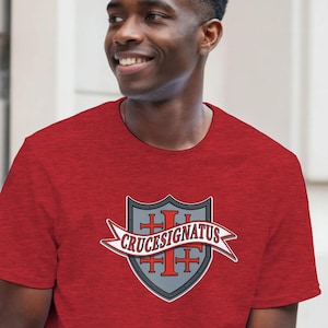 May include: A red short-sleeved t-shirt featuring a central graphic of a grey shield with a red cross emblem and a white banner overlay containing the black text CRUCESIGNATUS. The design is centred on the chest of the garment.