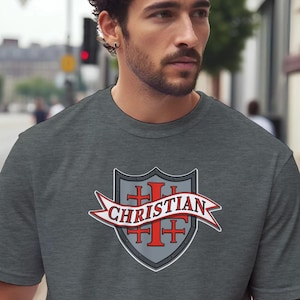 May include: A charcoal grey short-sleeve t-shirt featuring a central graphic of a shield with a red cross emblem. A white banner with a red outline is draped across the shield, displaying the word CHRISTIAN in bold, capitalised red letters.