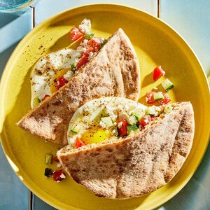 Può includere: Un piatto giallo contiene due metà di pane pita ripiene di uova fritte, pomodori a cubetti, cetrioli e formaggio sbriciolato. Le uova hanno tuorli giallo brillante. Il piatto è su una superficie azzurra.